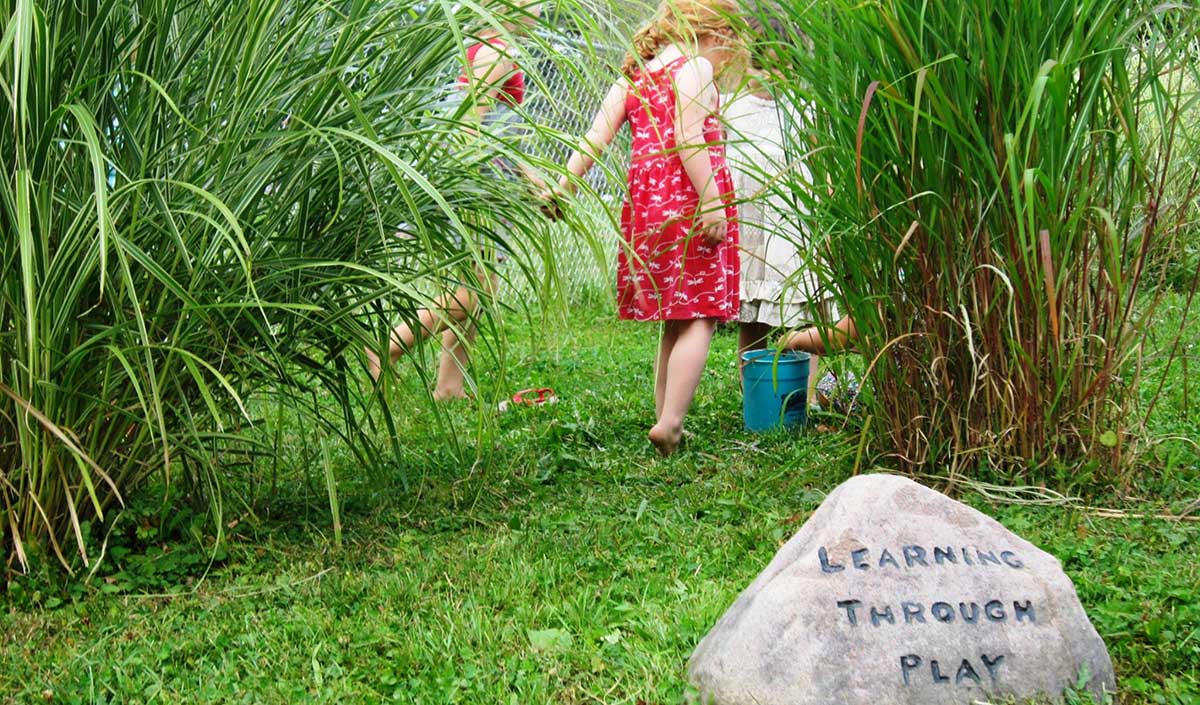 Children playing in tall grasses near a stone engraved with Learning Through Play