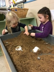 Two girls play at sensory table filled with sand.